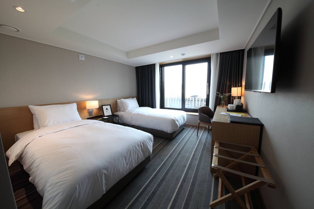 A hotel room with two beds, a desk, a chair, a TV, and a balcony view.