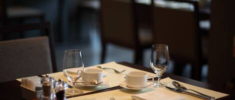 Una mesa de comedor dispuesta con tazas, platillos y cubiertos blancos, incluyendo un tenedor, cuchillo y cuchara.
