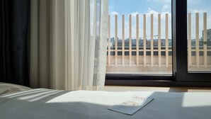 Habitació d'hotel amb una finestra gran, un llit i vista a un paisatge urbà.