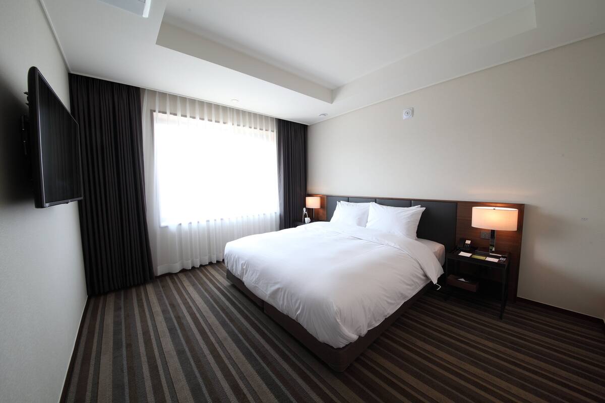 A hotel room with a large bed, two bedside tables with lamps, a flat-screen TV mounted on the wall, and a window with curtains.