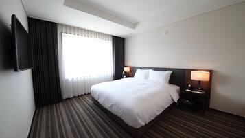 Hotellihuone, jossa on suuri sänky, kaksi yöpöytää valaisimineen, seinälle asennettu taulutelevisio ja ikkuna, jossa on verhot.