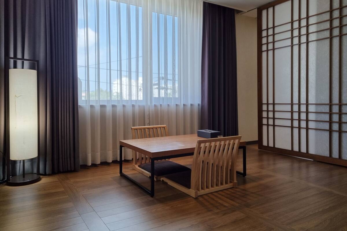 A room with a wooden table and chairs, a lamp, and a view through a window with sheer curtains.