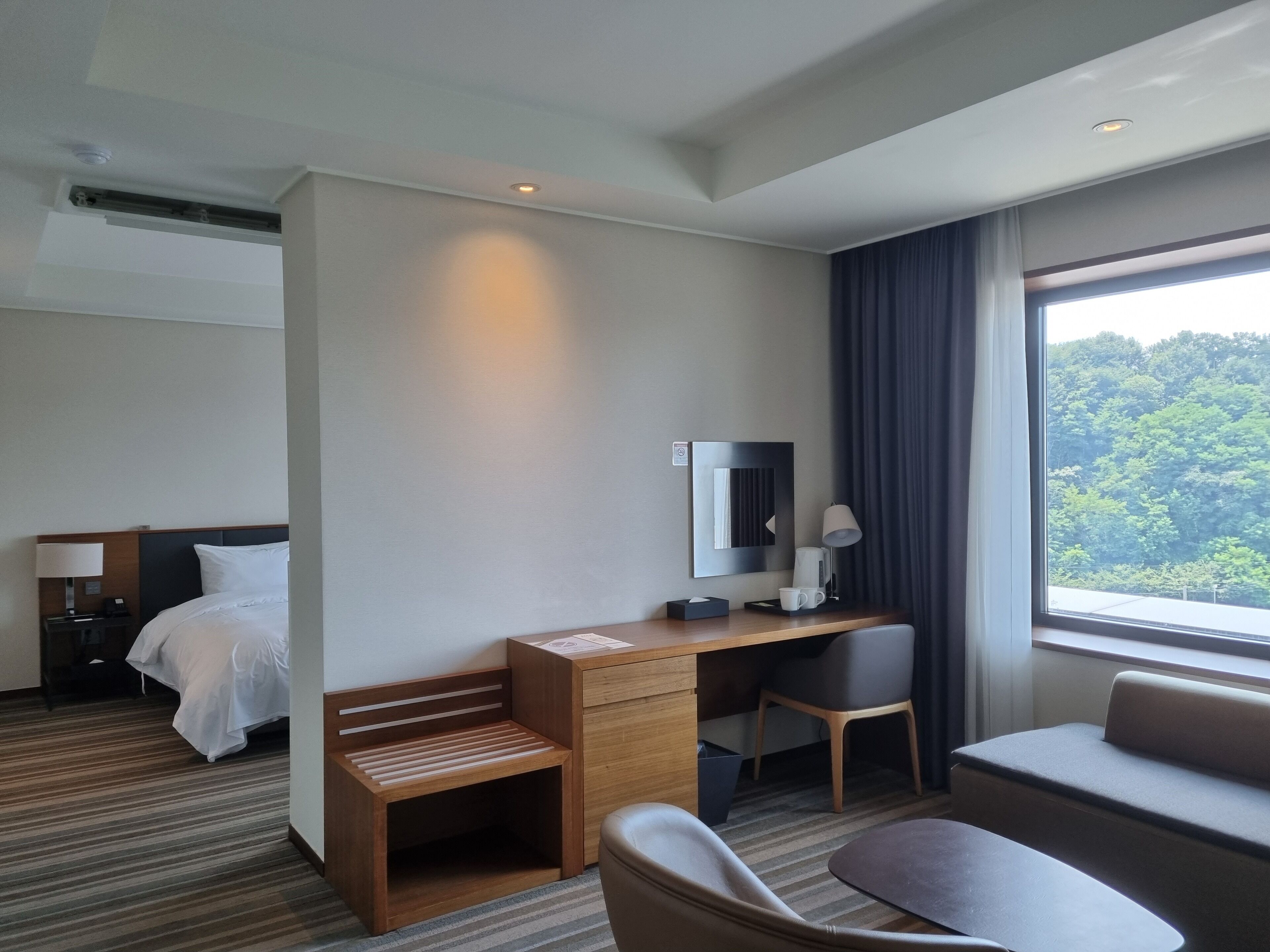 A hotel room with a bed, a desk with a chair, a small wooden bench, a bedside table with a lamp, and a large window with a view of trees.
