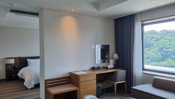 Una habitación de hotel con una cama, un escritorio con silla, un pequeño banco de madera, una mesita de noche con lámpara y un ventanal con vistas a árboles.