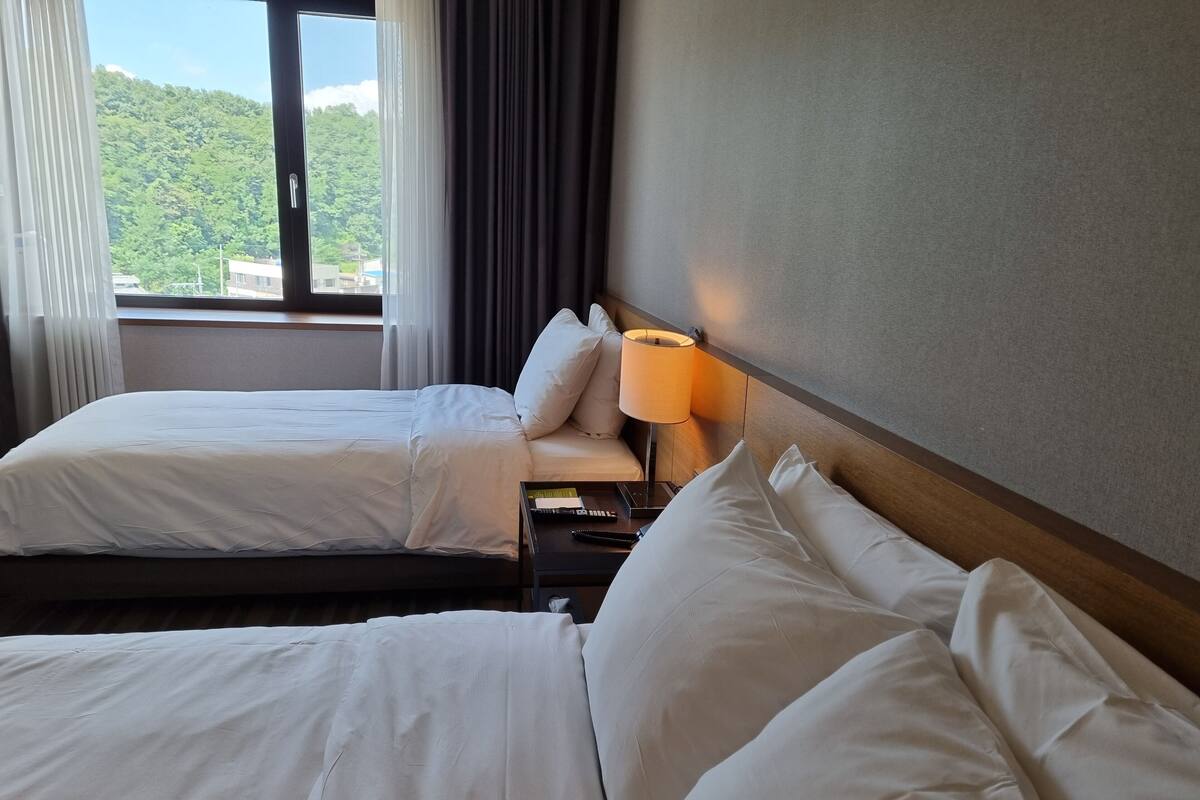 A hotel room with two beds, a nightstand with a lamp, and a view of a green landscape through the window.