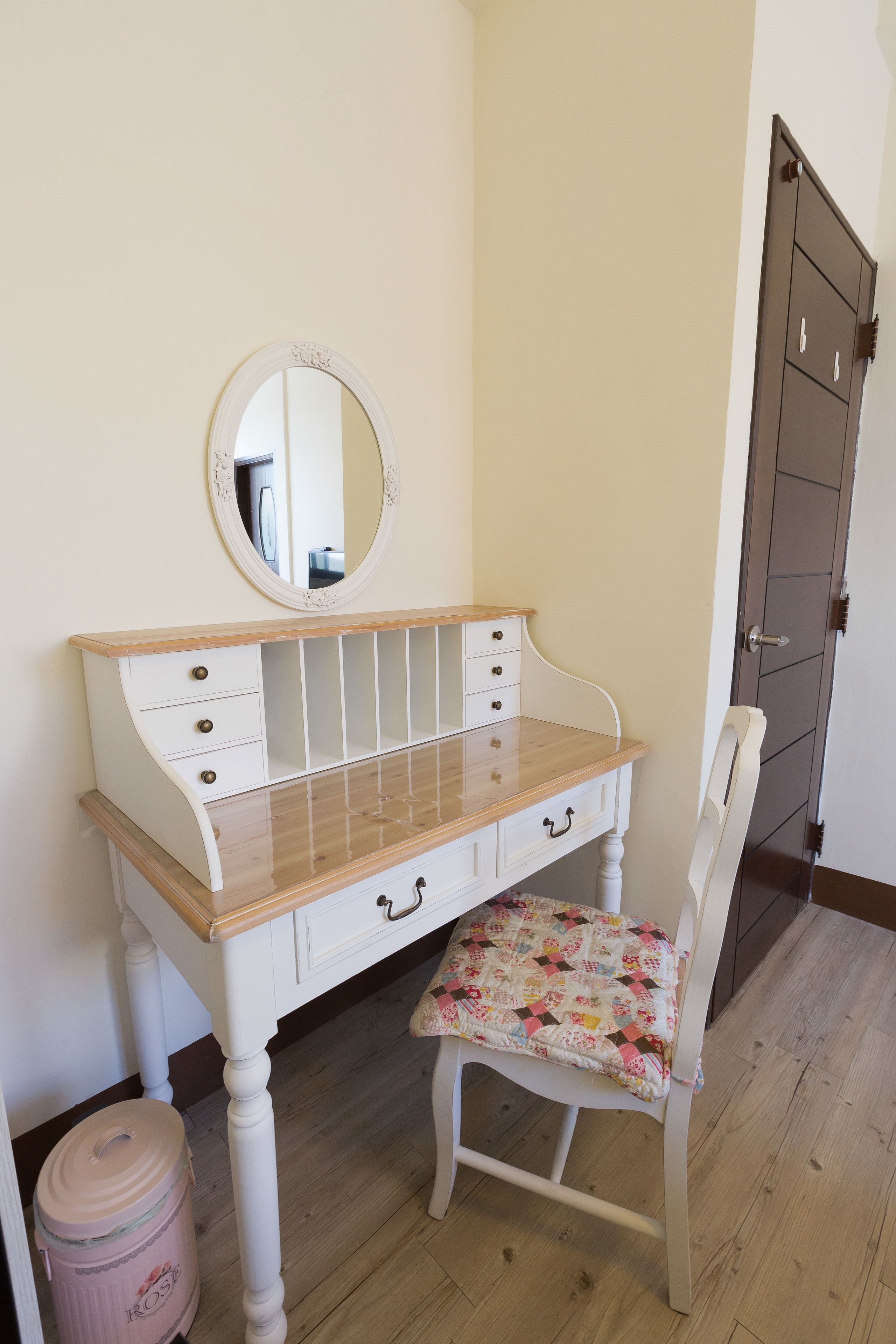 A white wooden desk with drawers and a mirror, a white chair with a floral cushion, and a wooden floor.