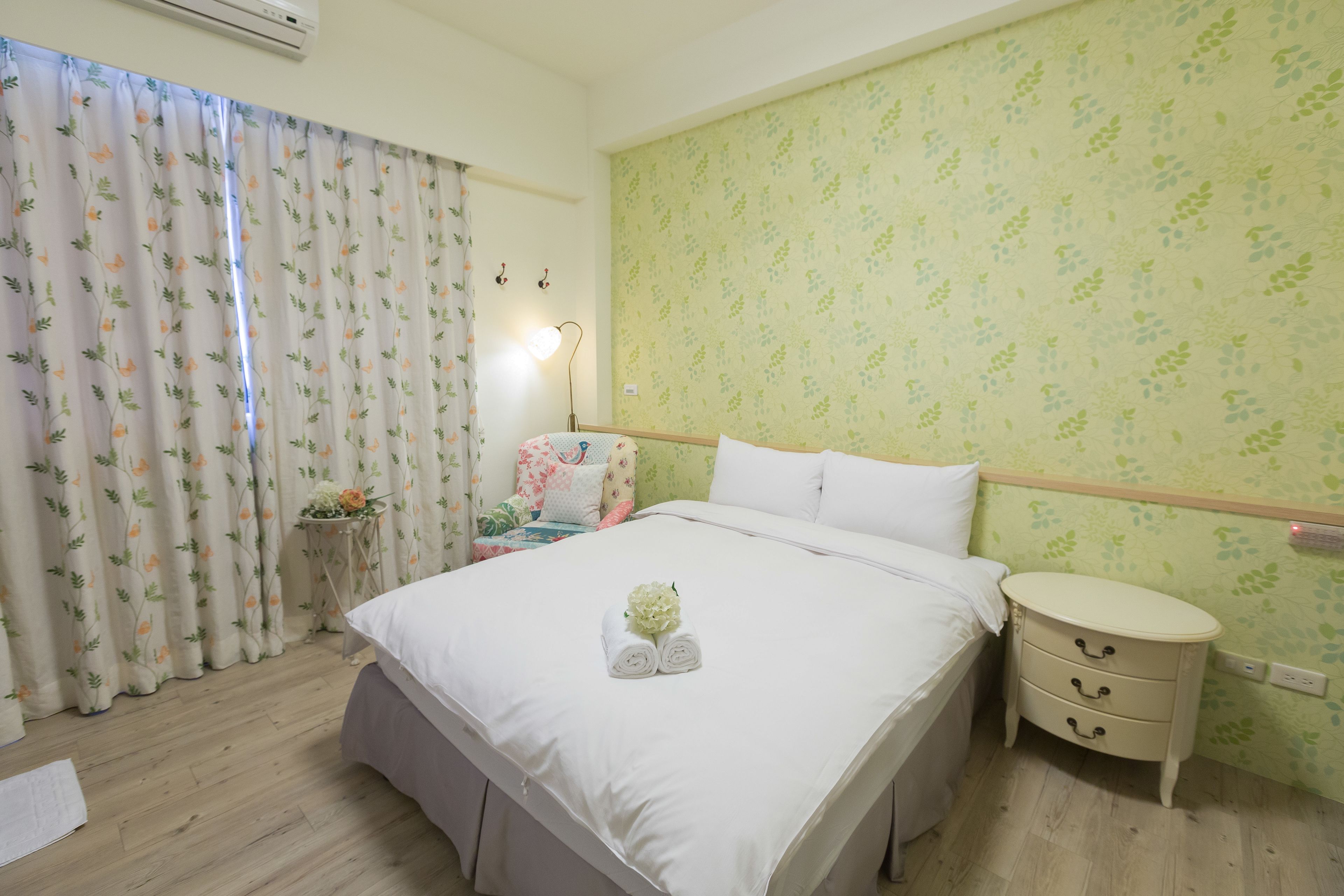 Habitación de hotel con una cama grande, una mesita de noche con cajones, una pequeña mesa con lámpara y una ventana con cortinas florales.