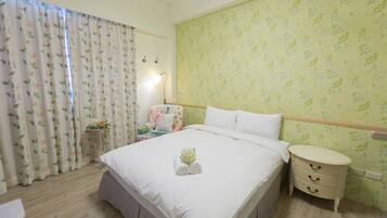 Habitación de hotel con una cama grande, una mesita de noche con cajones, una pequeña mesa con lámpara y una ventana con cortinas florales.