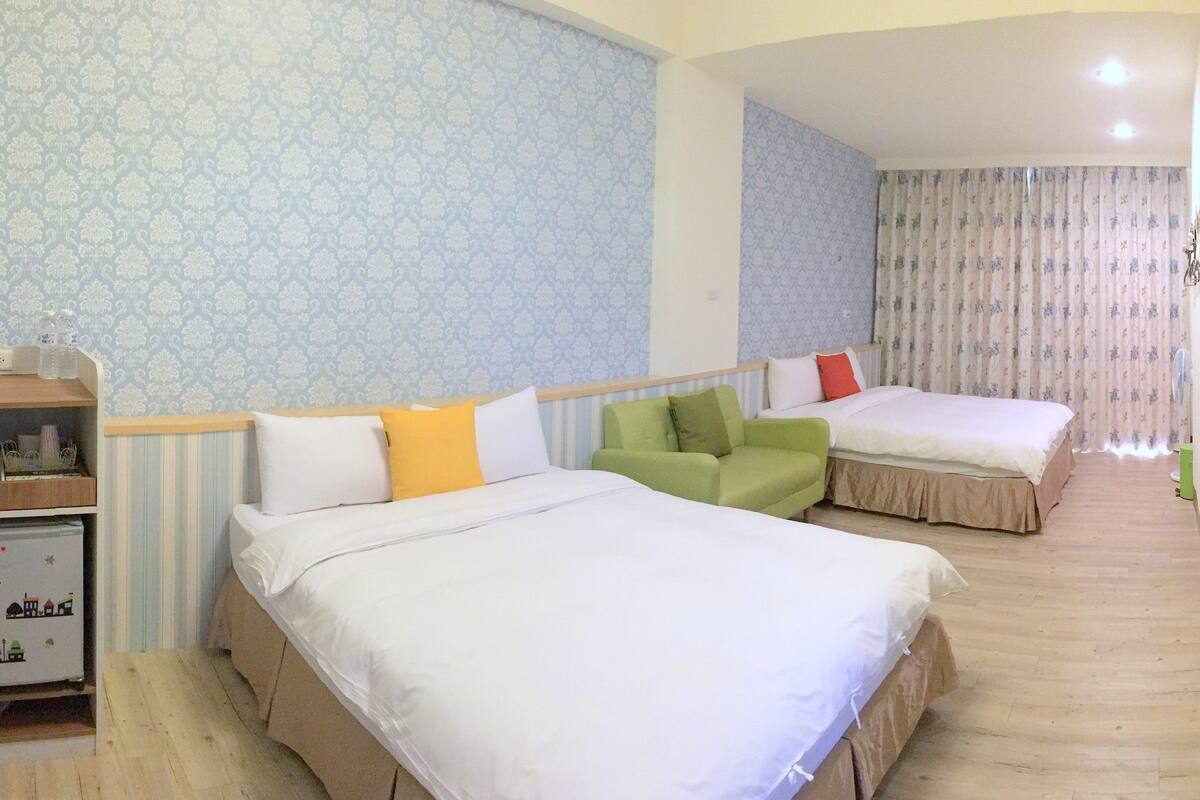 A hotel room with two beds, a small sofa, a desk with a kettle, and a shelf with toiletries.