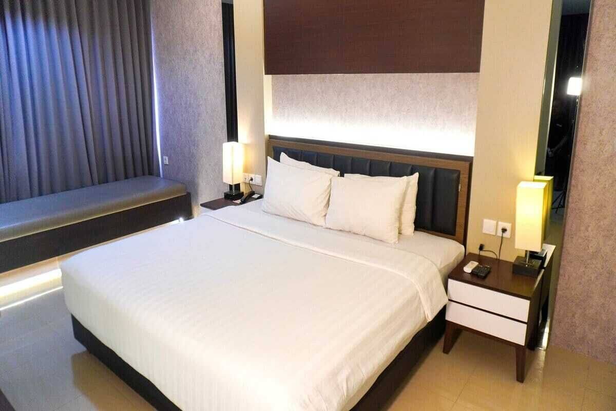 A hotel room with a large bed, two bedside tables with lamps, a headboard, and a window with curtains.