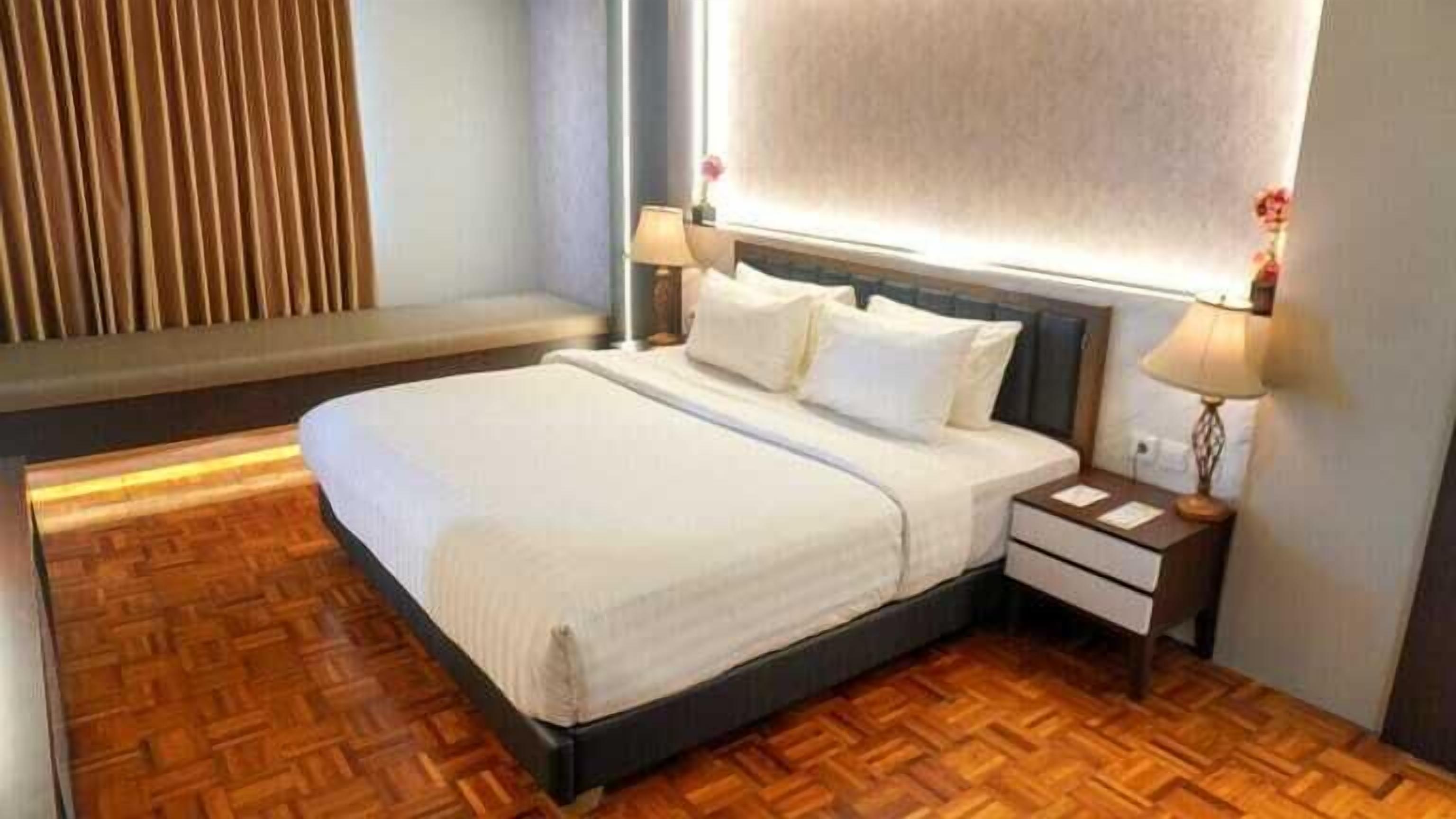 A neatly made bed in a hotel room with a wooden floor, two bedside tables with lamps, and a large window with blinds.