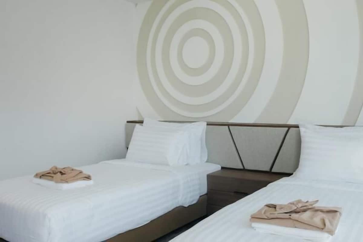 A hotel room with two single beds, each with a white bedspread and a folded beige towel. The headboard has a geometric design, and the wall features a circular pattern.