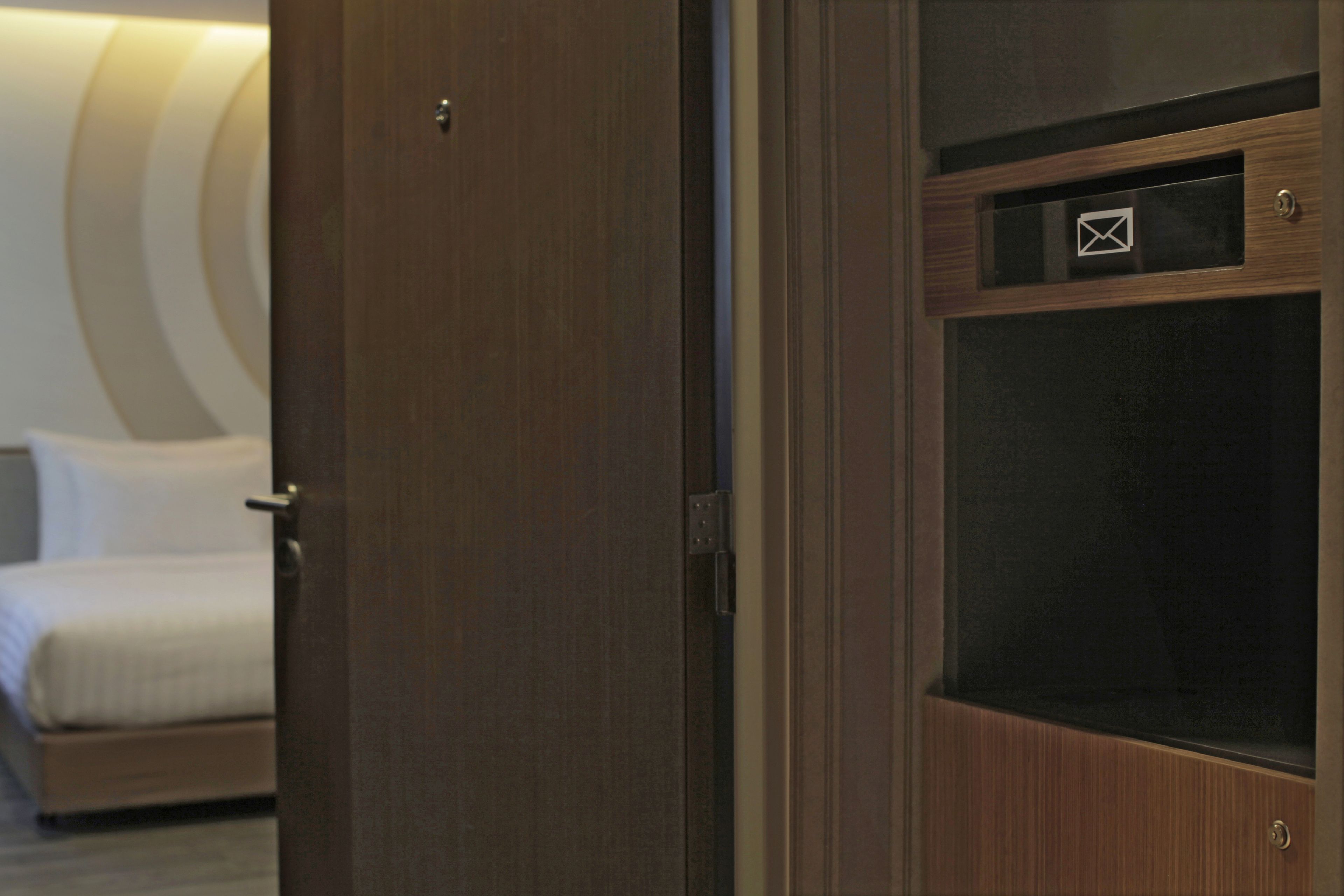 A hotel room with a wooden door, a bed with white bedding, and a small window with an envelope symbol.
