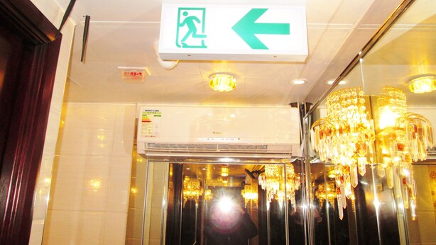 An elevator interior with a green emergency exit sign, a glass door, and a chandelier. - New Reliance Inn (Kowloon)