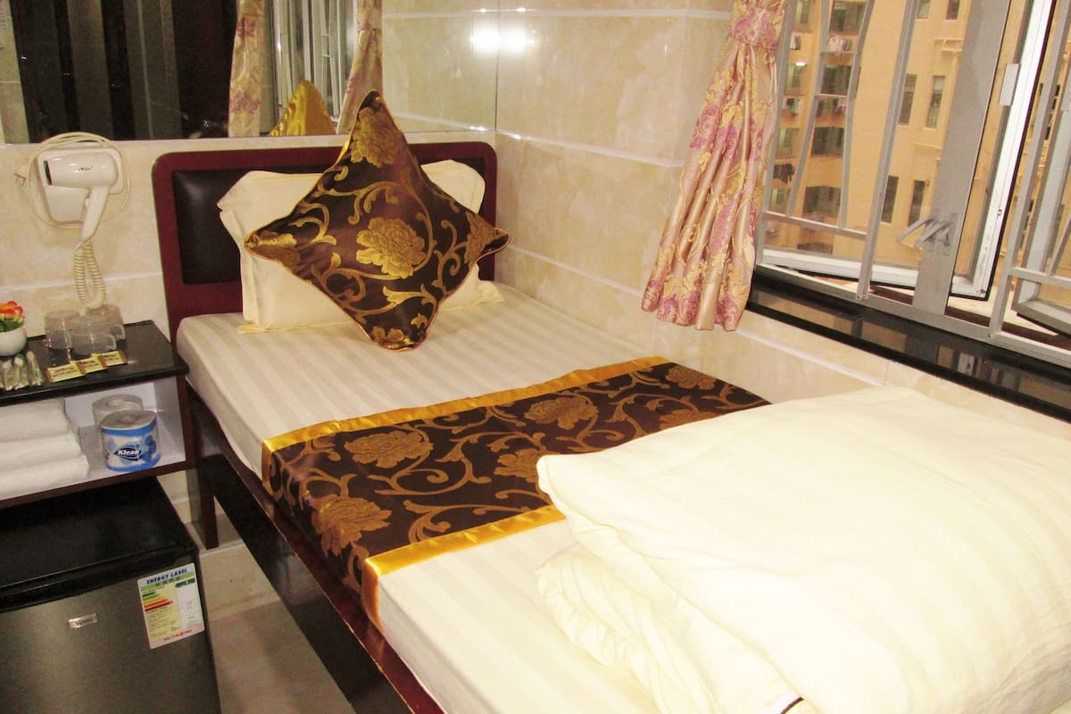 A hotel room with a bed, a bedside table with a hairdryer, a bottle of lotion, and a small refrigerator.