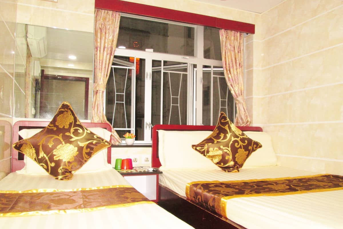 A hotel room with two beds, each with a decorative pillow, a window with curtains, and a small table with a vase and cups.