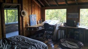 Una cabaña rústica con interior de madera, un escritorio con computadora, una cama y un ventanal con vistas a árboles.