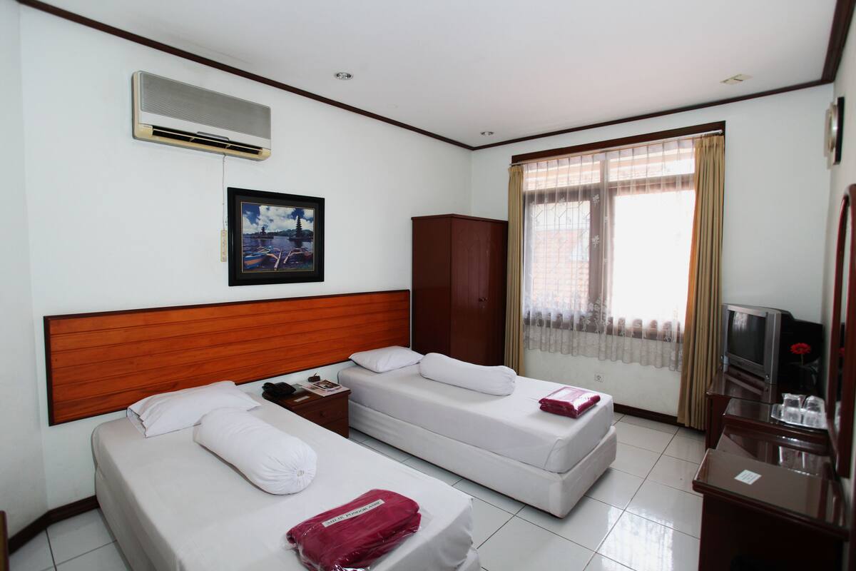 Habitación de hotel con dos camas individuales, un cabecero de madera, un televisor, un armario y una ventana con cortinas.