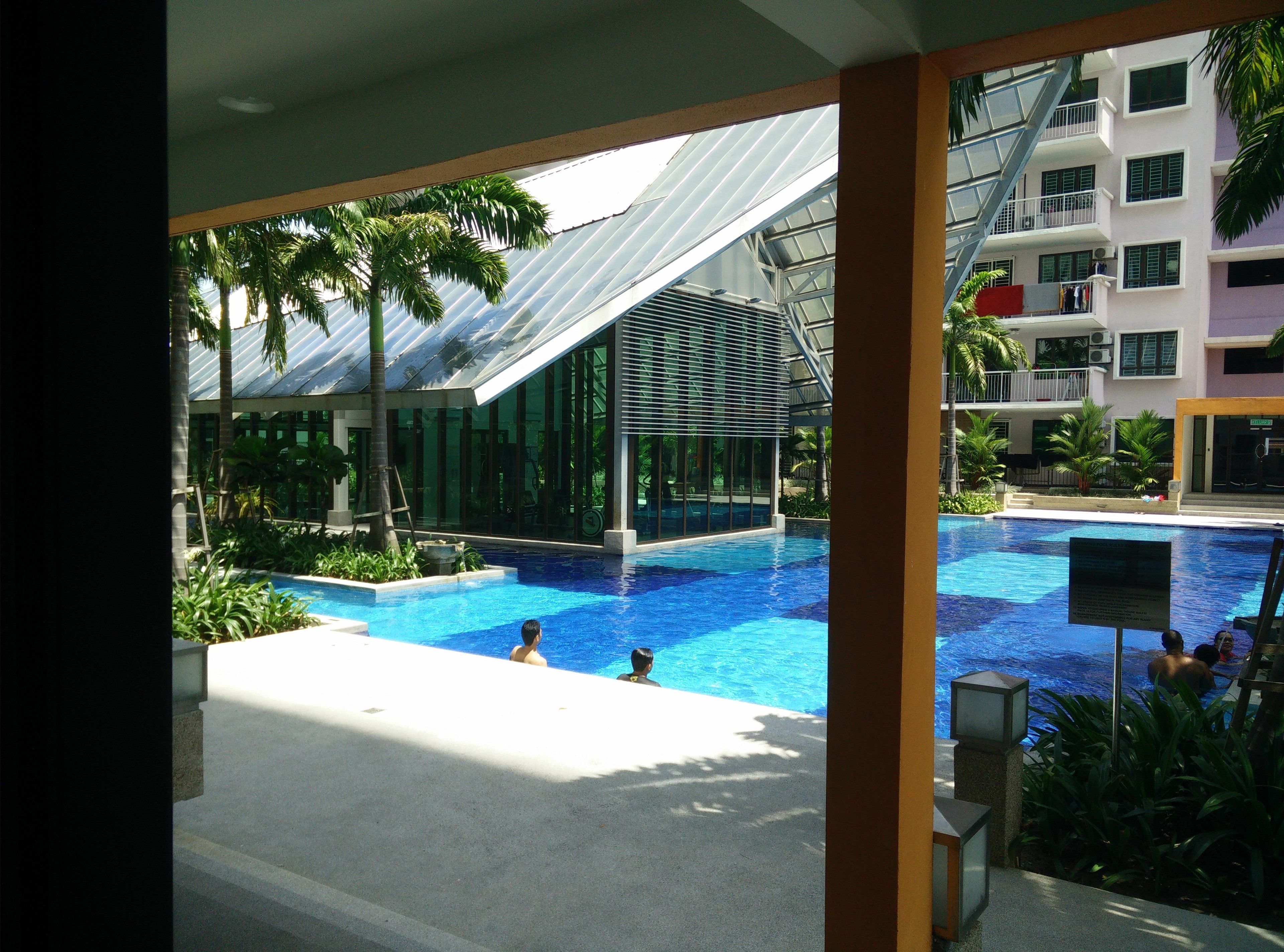 A swimming pool with people, surrounded by palm trees and modern buildings.