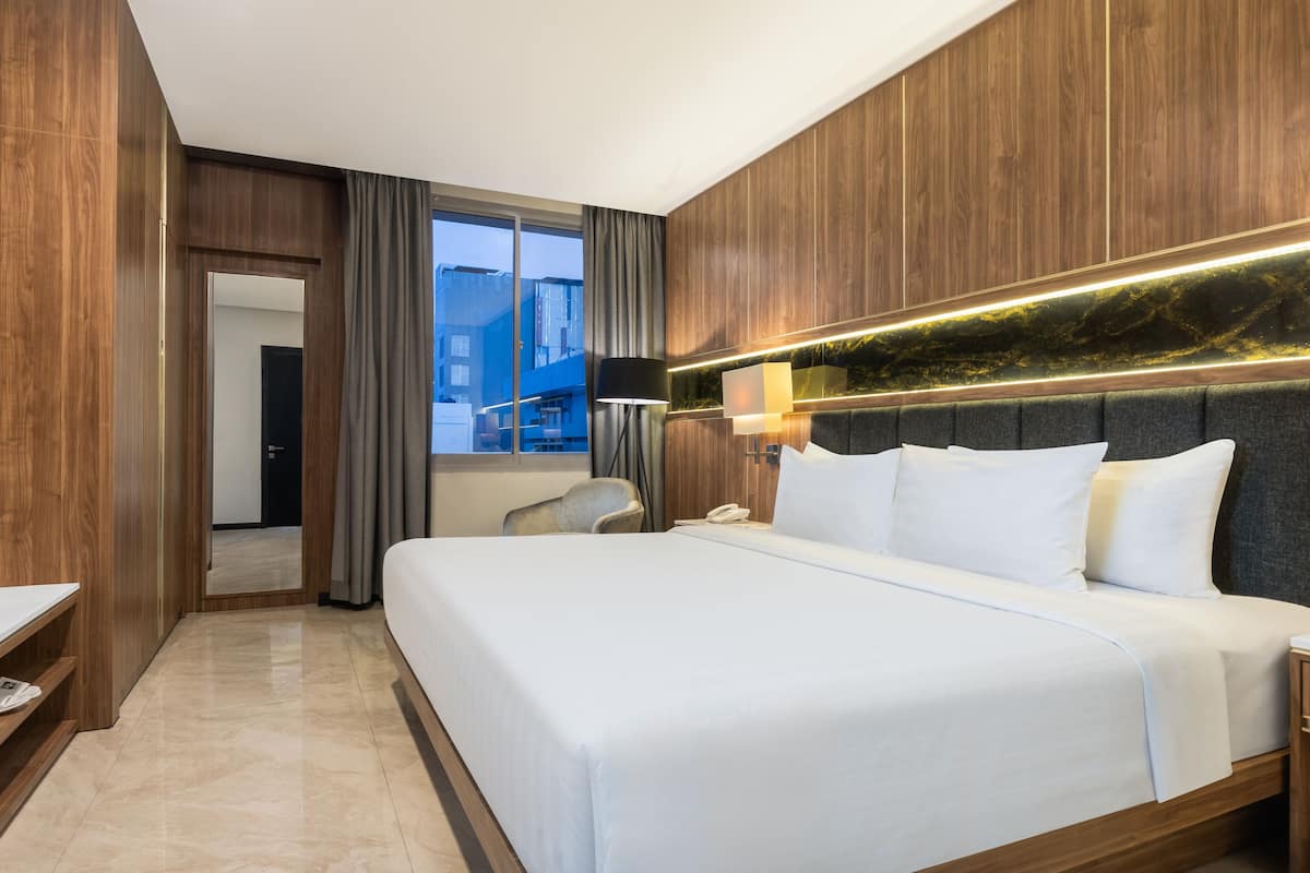 Ein Hotelzimmer mit einem großen Bett, einem Schreibtisch, einem Sessel und Blick auf die Stadt durch das Fenster.