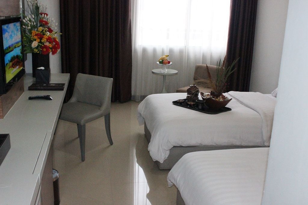 Habitación de hotel con dos camas, un escritorio con televisor, una silla, una mesita con frutero y un florero.