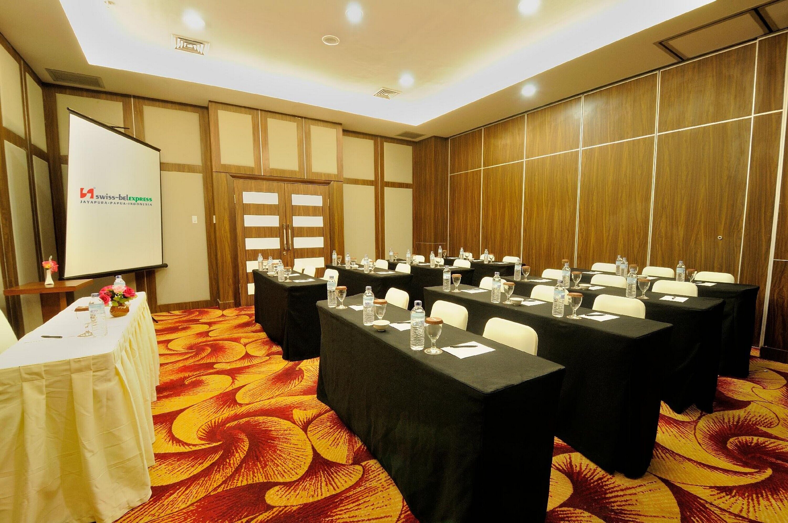 Uma sala de conferências com mesas preparadas para uma reunião, uma tela de projeção e um arranjo floral sobre uma mesa.