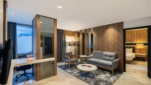 Ein modernes Hotelzimmer mit großem Schreibtisch, einem grauen Sofa, einem kleinen runden Tisch und einem großen Spiegel.