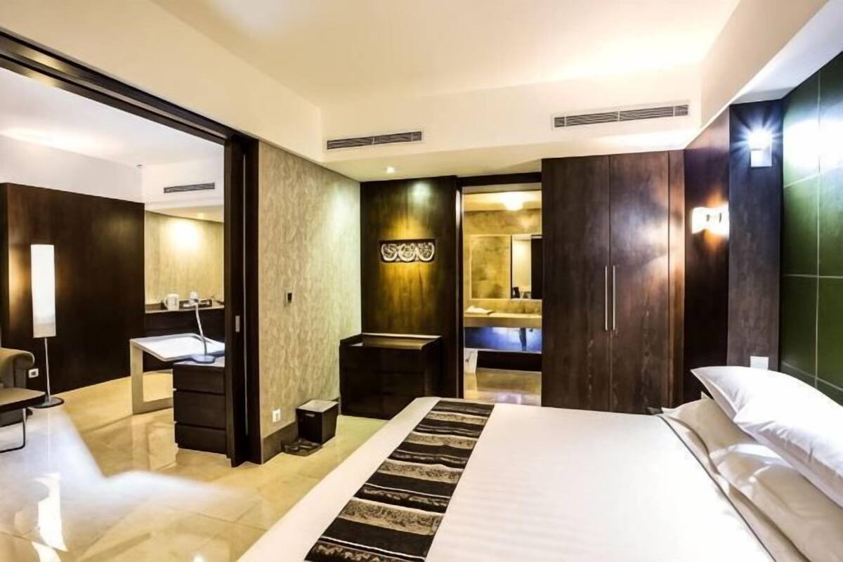 A hotel room with a large bed, a desk with a chair, a mirror, and a bathroom visible through an open door.