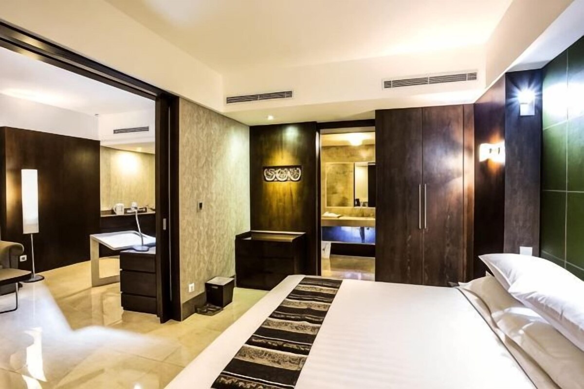 A hotel room with a large bed, a desk with a chair, a mirror, and a bathroom visible through an open door.