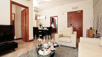 A living room with a flat-screen TV, a sofa, a coffee table with a floral arrangement, and a dining area with a table set for four.