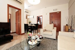 A living room with a flat-screen TV, a sofa, a coffee table with a floral arrangement, and a dining area with a table set for four.