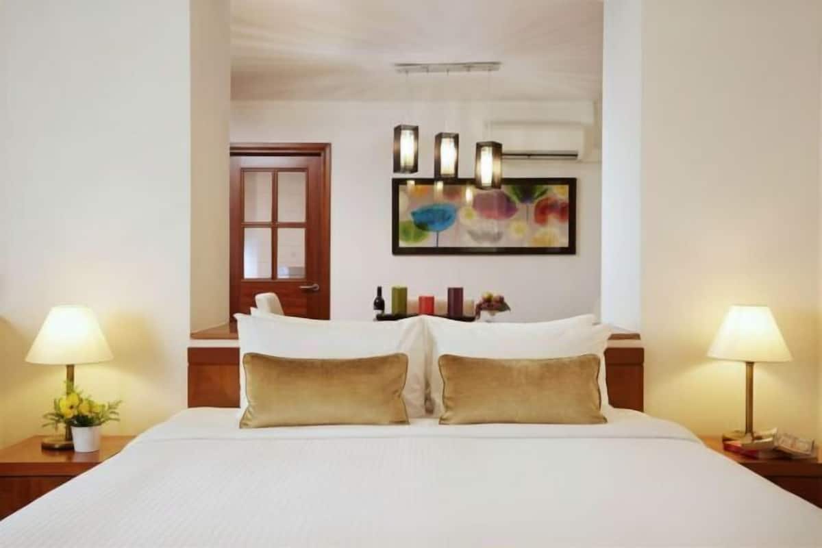 A hotel room with a large bed, two bedside lamps, a door, a wall-mounted artwork, and a small table with a plant.