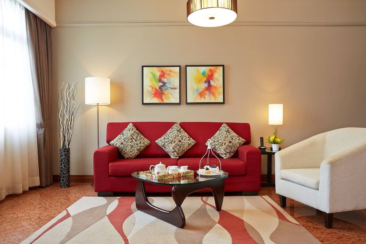 A living room with a red sofa, two framed abstract paintings, a glass coffee table with tea sets, a white armchair, and a patterned rug.