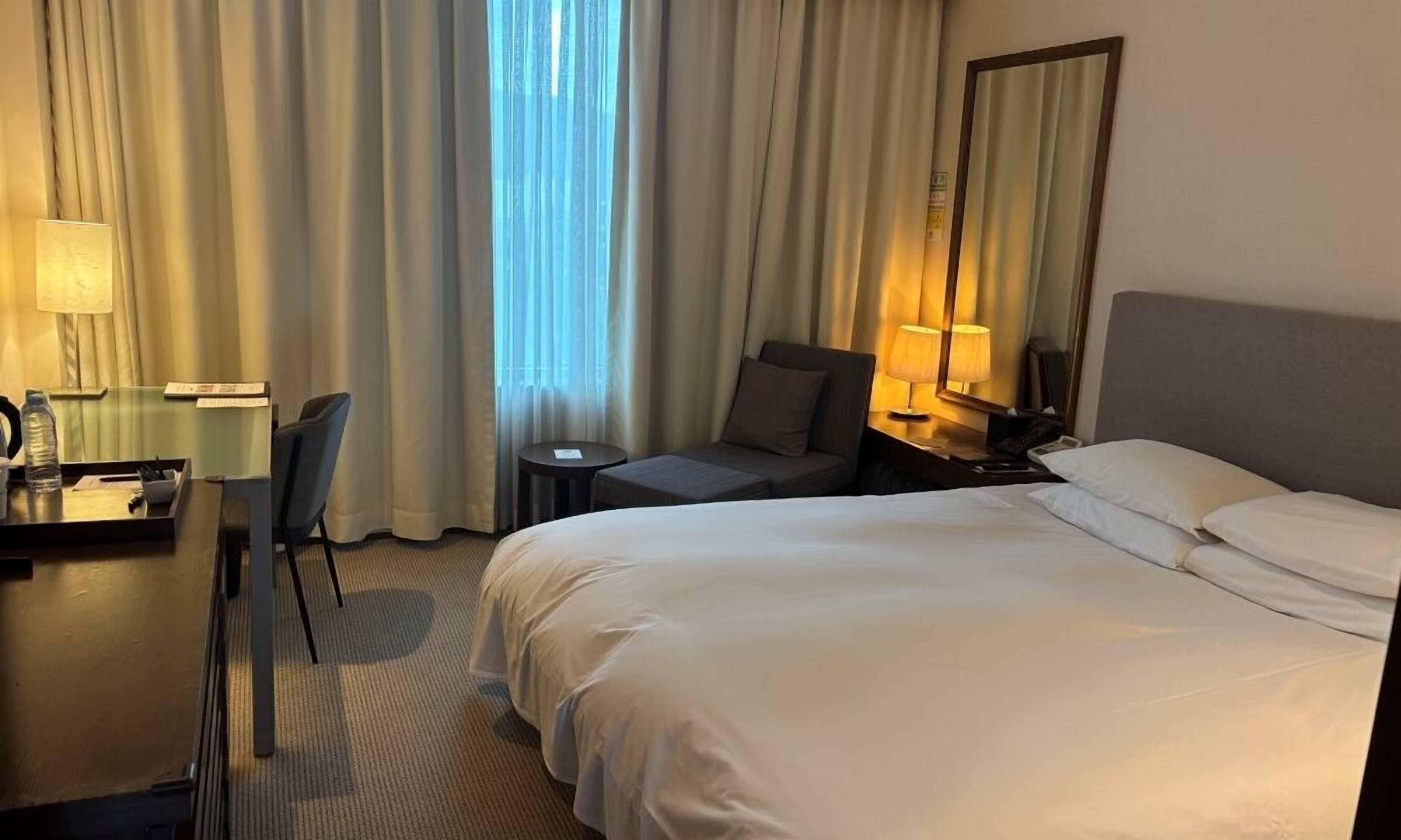 A hotel room with a bed, a desk, a chair, a nightstand, and a lamp.