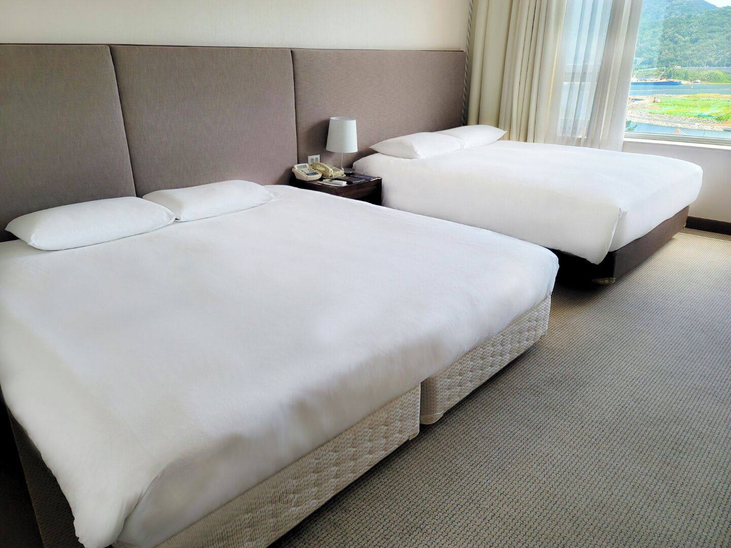 A hotel room with two beds, a nightstand with a lamp, and a view of a lake through the window.