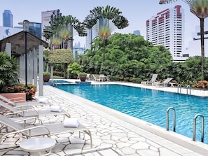 A pool area with lounge chairs and a small pavilion, surrounded by lush greenery and tall buildings in the background.
