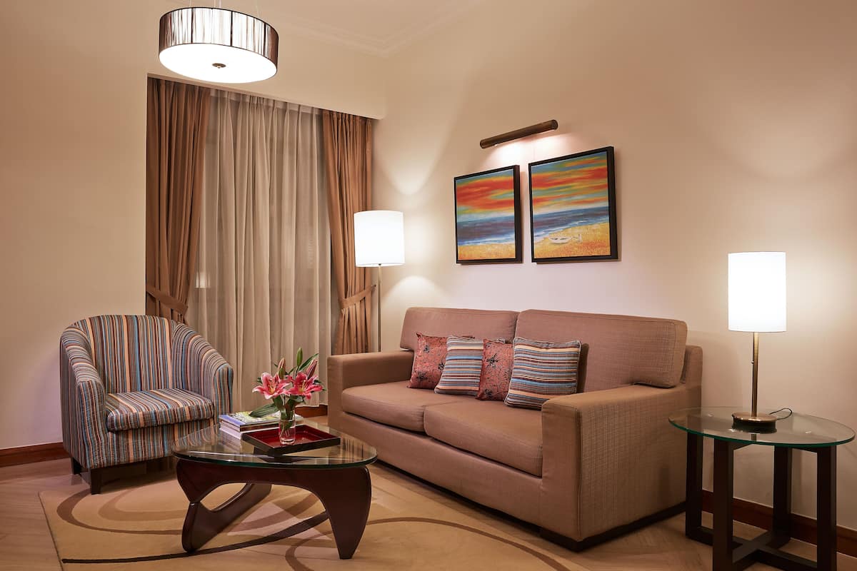 A living room with a striped armchair, a sofa with patterned cushions, a glass coffee table, and two wall-mounted lamps.