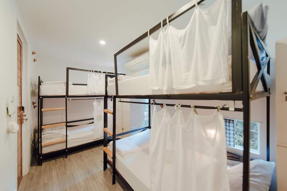 A dormitory room with bunk beds, wooden flooring, and a window with a view of greenery.