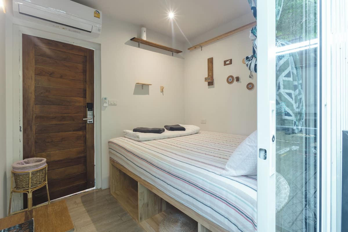A bedroom with a wooden door, a bed with striped bedding, a small wooden stool, and a window with a view of greenery.