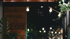 En kat ligger på jorden i et svagt oplyst udendørs område med et træskilt, der siger "WELCOME TO THE YARD FAMILY".