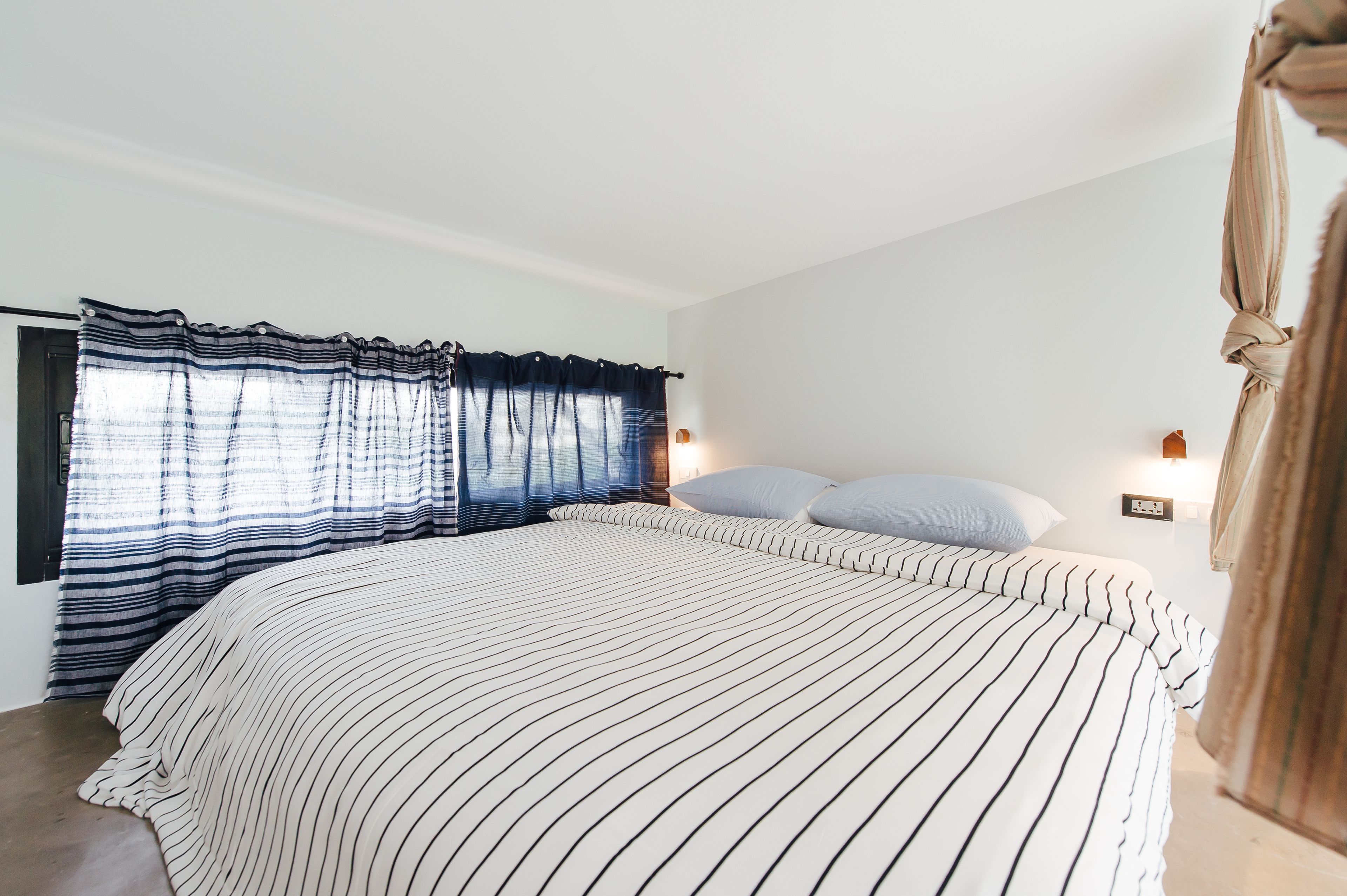 A bedroom with a large bed, striped bedding, a window with curtains, and a wall-mounted light.