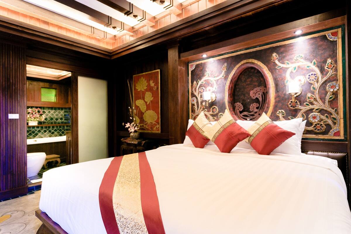 Ein Hotelzimmer mit einem großen Bett, einem Holzkopfteil mit aufwendigen Verzierungen, einem kleinen Tisch mit Blumenvase und einem im Hintergrund sichtbaren Badezimmer.