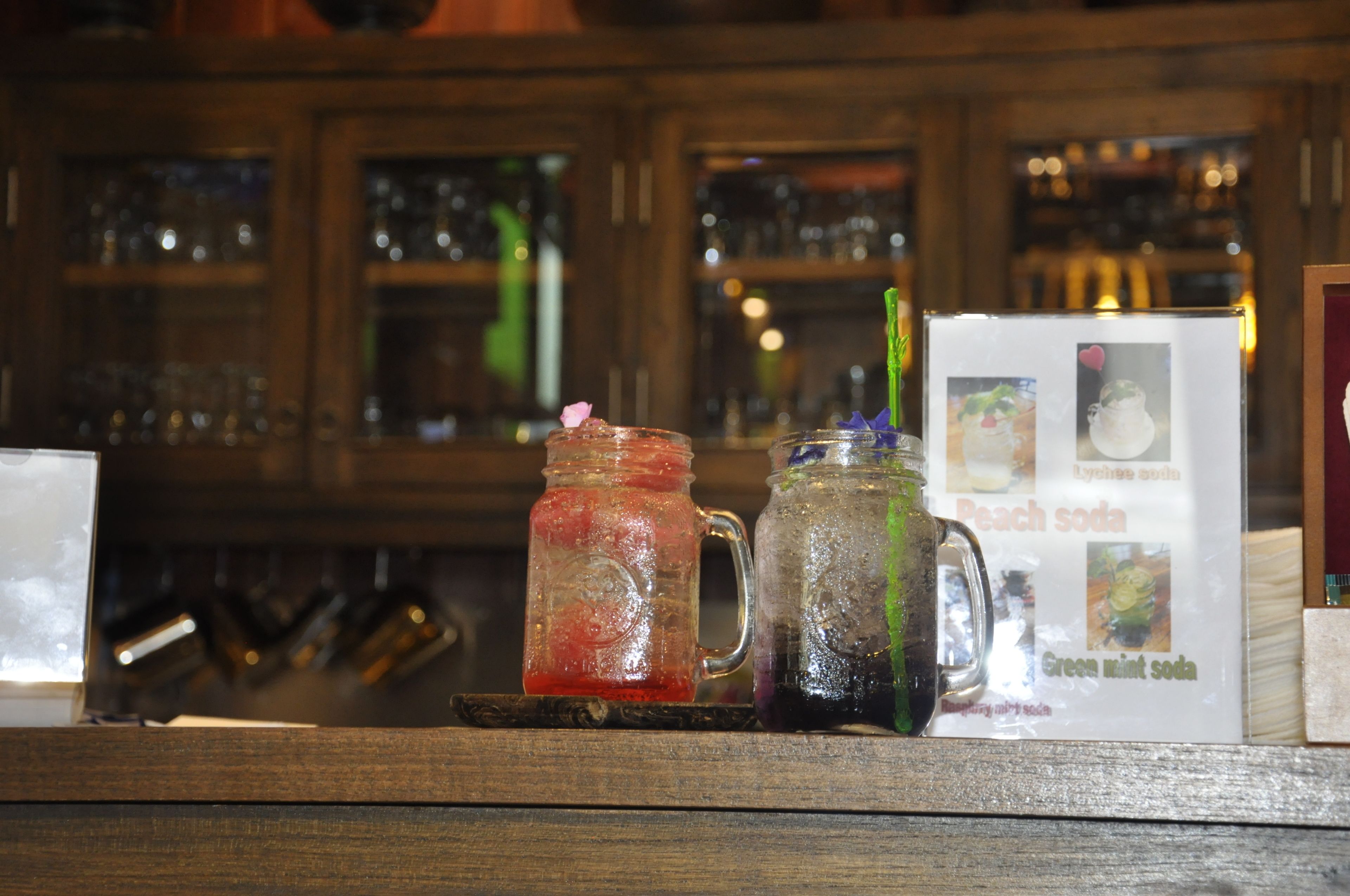 Zwei Gläser mit Getränken auf einer Holzablage, dahinter eine Speisekarte und im Hintergrund eine Bar.
