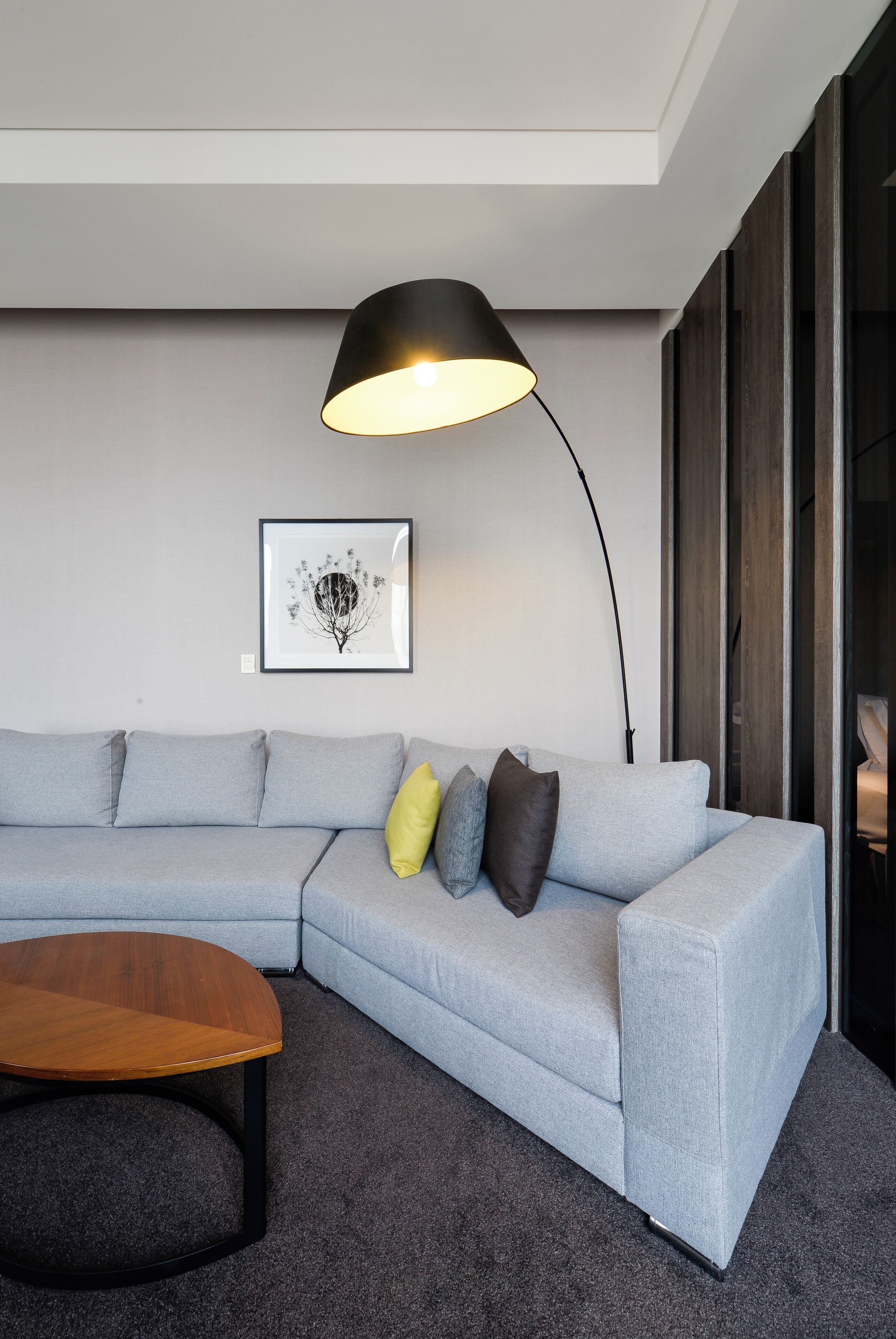 Ein modernes Wohnzimmer mit einem grauen Ecksofa, einem Holztisch, einer schwarzen Stehlampe und einem gerahmten Kunstwerk an der Wand.