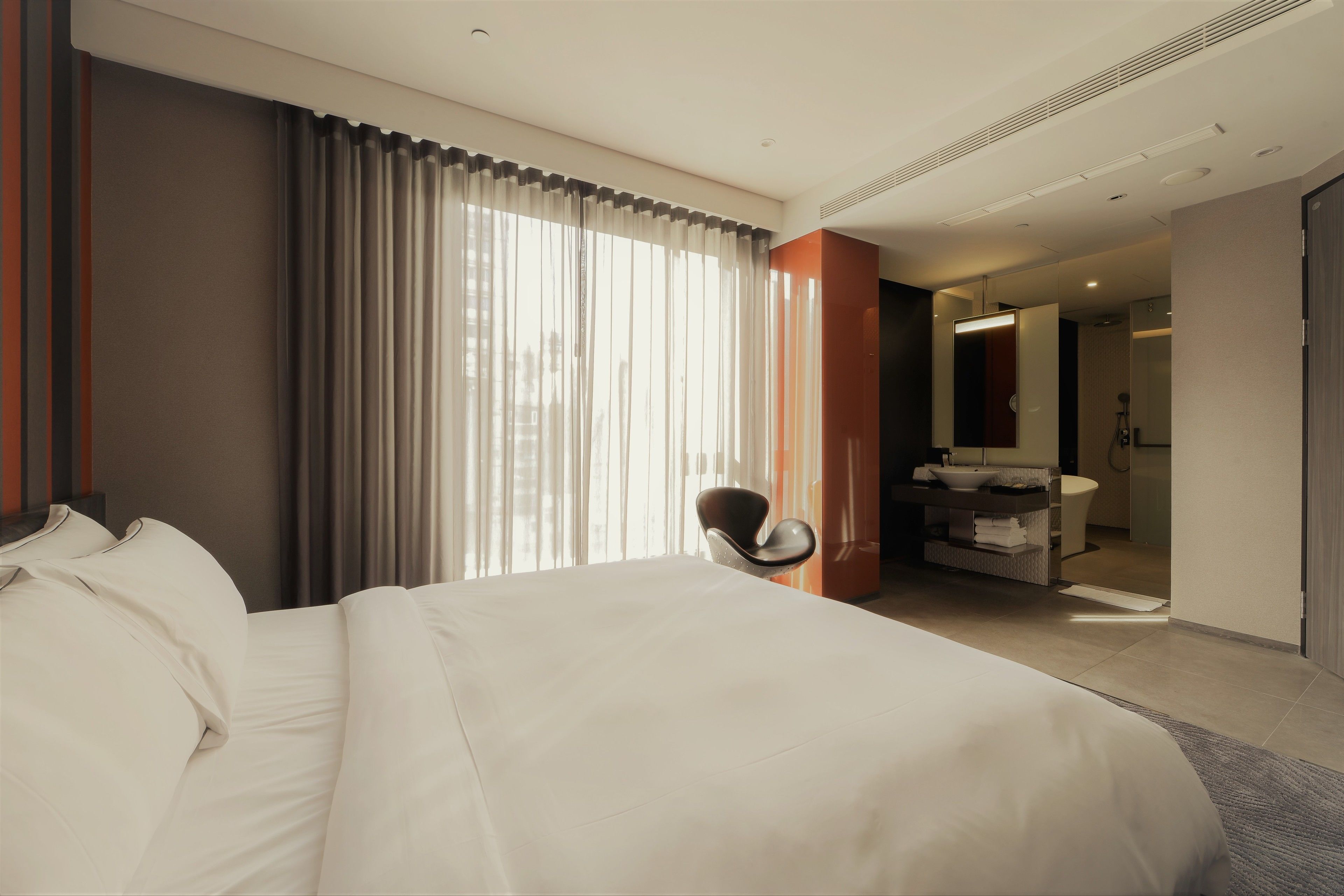 Ein modernes Hotelzimmer mit einem großen Bett, einem Sessel, einem Badezimmer mit Waschbecken und Spiegel sowie Blick auf die Stadt durch große Fenster.
