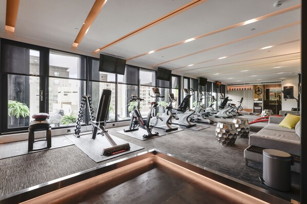 A modern gym with various exercise equipment, large windows, and a seating area with a sofa and coffee table.