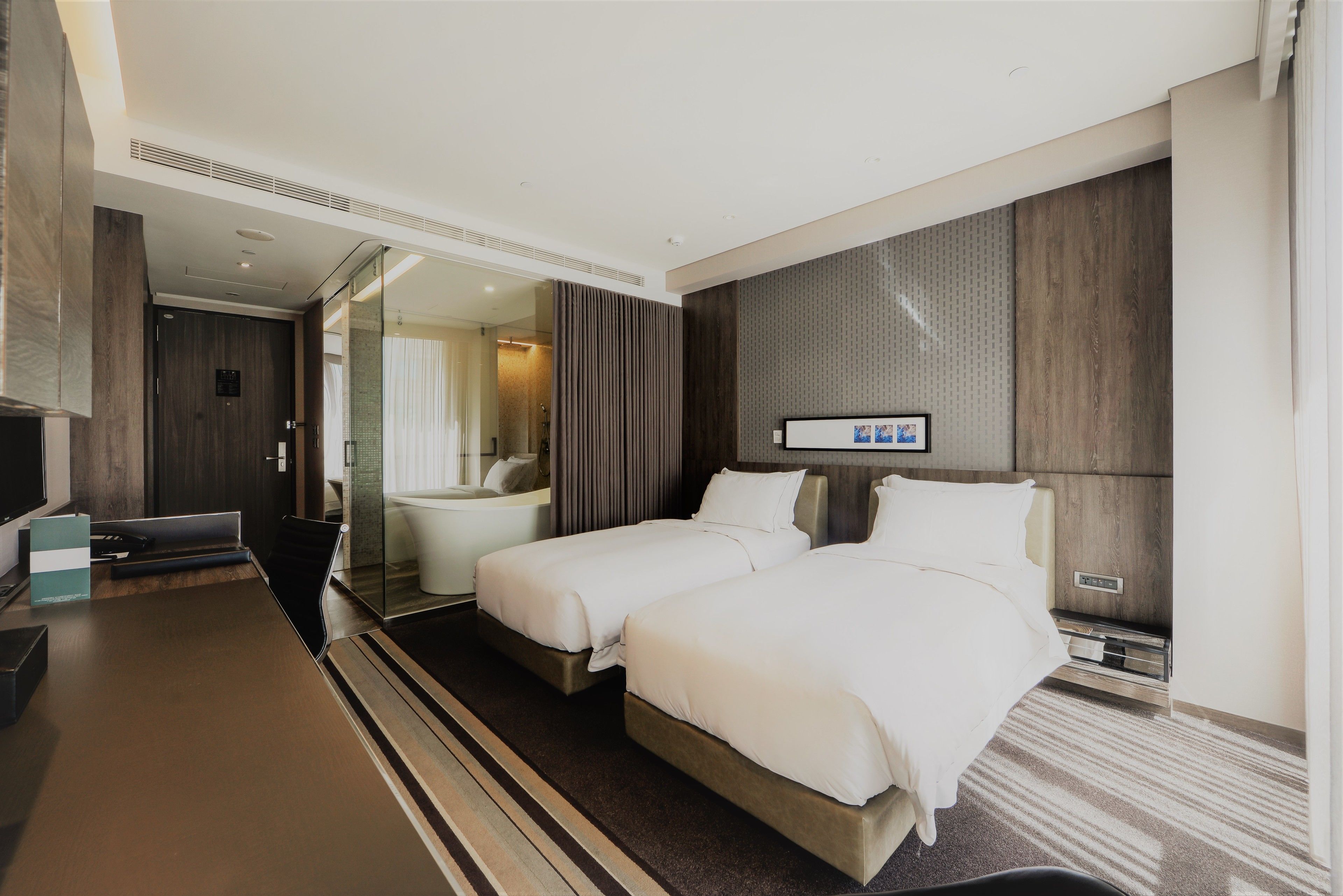 A modern hotel room with two beds, a large flat-screen TV, a desk, and a bathroom with a glass shower.