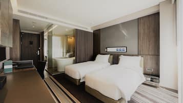 A modern hotel room with two beds, a large flat-screen TV, a desk, and a bathroom with a glass shower.