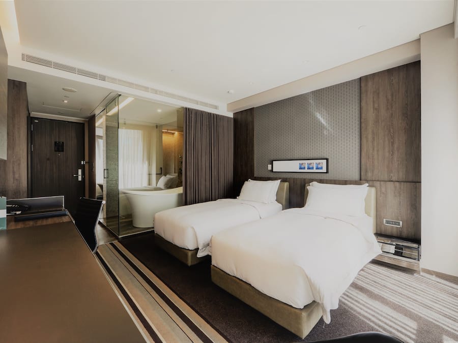 A modern hotel room with two beds, a large flat-screen TV, a desk, and a bathroom with a glass shower.