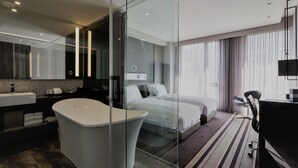 Habitación de hotel moderna con ducha de vidrio, una bañera grande, cama doble, escritorio con silla y un ventanal con cortinas.